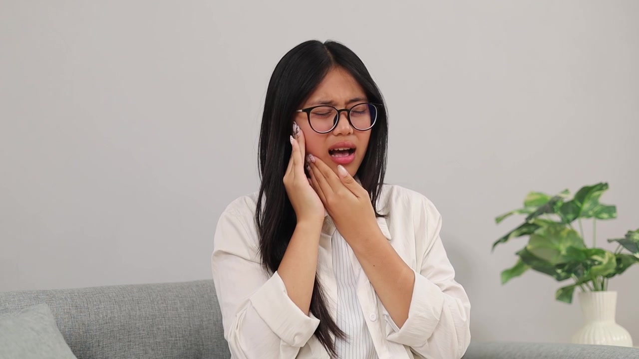 Young Asian woman sitting on sofa while touching her sore jaw and suffering from stubbing toothache