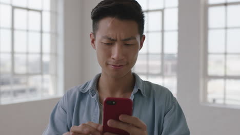 close-up-portrait-of-young-asian-man-student-texting-browsing-social-media-using-smartphone-mobile-technology-in-empty-apartment-room