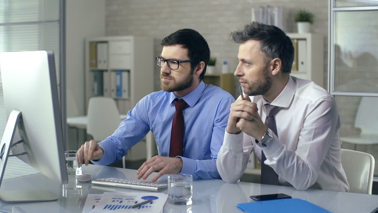 Two Businessmen In Shirts And Ties Talking About A Project Looking At The Computer Screen Sitting At A Table In The Office