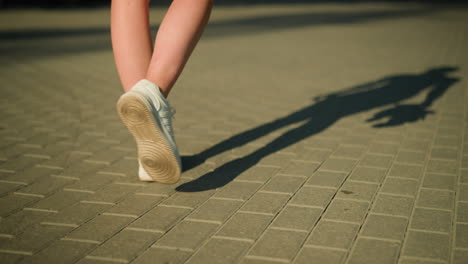 back view of individual wearing white canvas shoes walking on interlocked path with shadow cast on the ground, sunlight illuminates legs and pathway