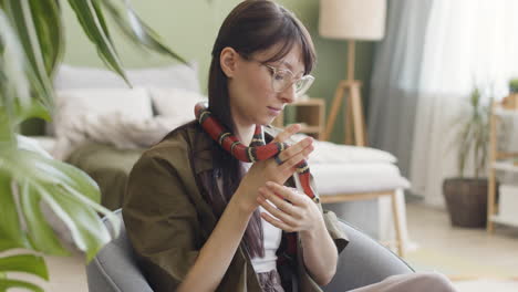 Smiling-Young-Woman-With-Eyeglasses-Holding-Her-Pet-Snake-While-Sitting-In-An-Armchair-At-Home