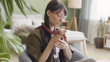 smiling young woman with eyeglasses holding her pet snake while sitting in an armchair at home