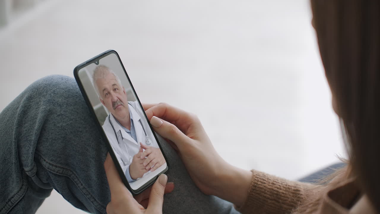 Female medical assistant wears white coat headset video calling distant patient on smartphone. Doctor talking to client using virtual chat telephone app. Telemedicine remote healthcare services
