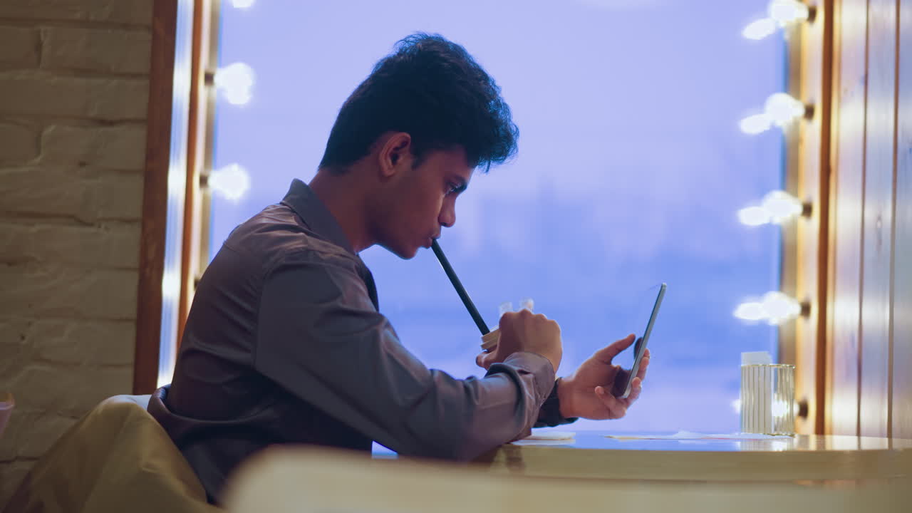 Young man sitting alone at table in cozy cafe, sipping drink through straw while looking at smartphone in hand, surrounded by warm lights and soft indoor ambiance, showing digital lifestyle and solitude