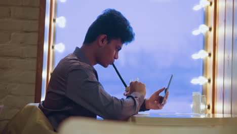 young man sitting alone at table in cozy cafe, sipping drink through straw while looking at smartphone in hand, surrounded by warm lights and soft indoor ambiance, showing digital lifestyle and solitude