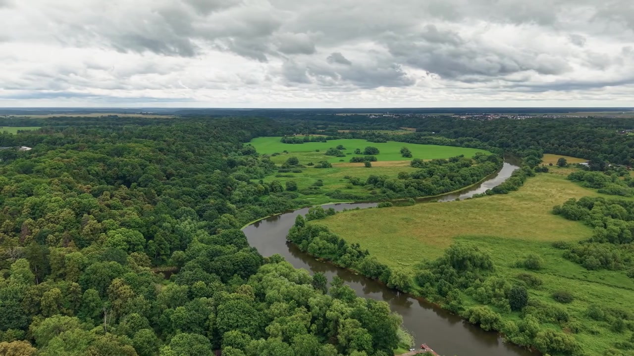 Vídeo de drone deslumbrante captura um rio sinuoso serpenteando por prados verdejantes e floresta densa sob nuvens dramáticas, exibindo uma rica vegetação e natureza selvagem intocada vista de cima.