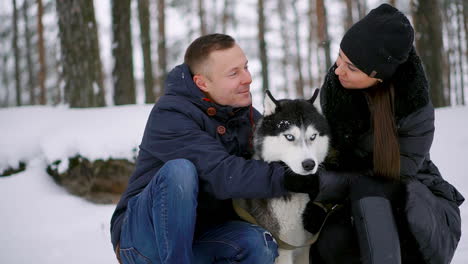 a man and a woman sitting hugging a dog siberian husky in the winter forest smiling and looking at each other and at the camera. slow motion