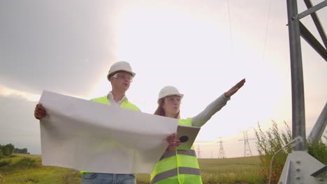 Two-electricians-work-together-standing-in-the-field-near-electricity-transmission-line-in-helmets.-Two-electricians-work-together-standing-in-the-field-near-with-power-transmission-towers.-Eco-friendly-fuel