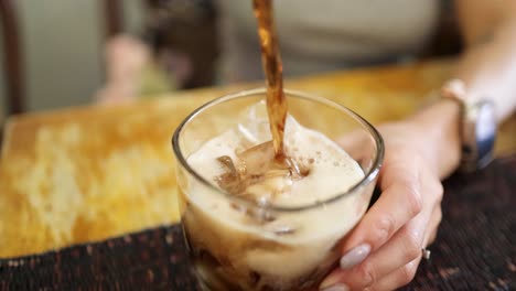 a person pours cola from a can into a glass filled with ice, creating foam. shot indoors with natural lighting and a steady, close-up perspective