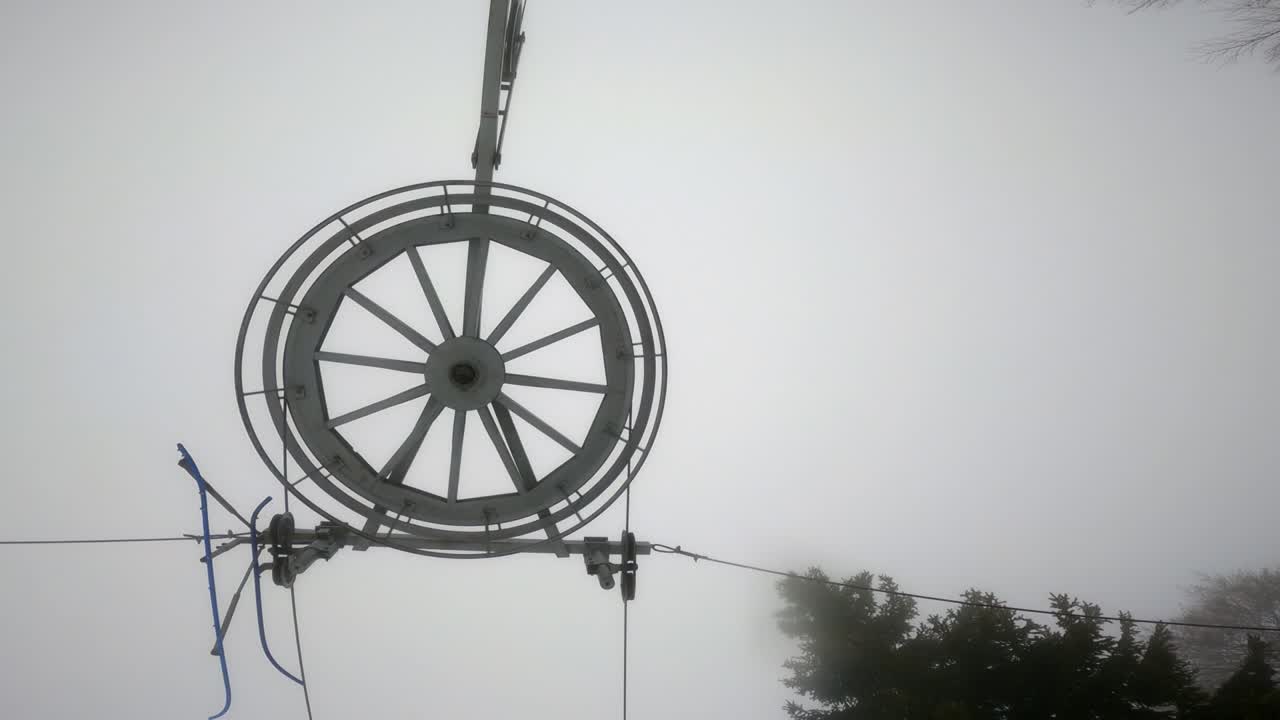 Ski lift pulley, wires, and wheel against a cloudy and foggy sky