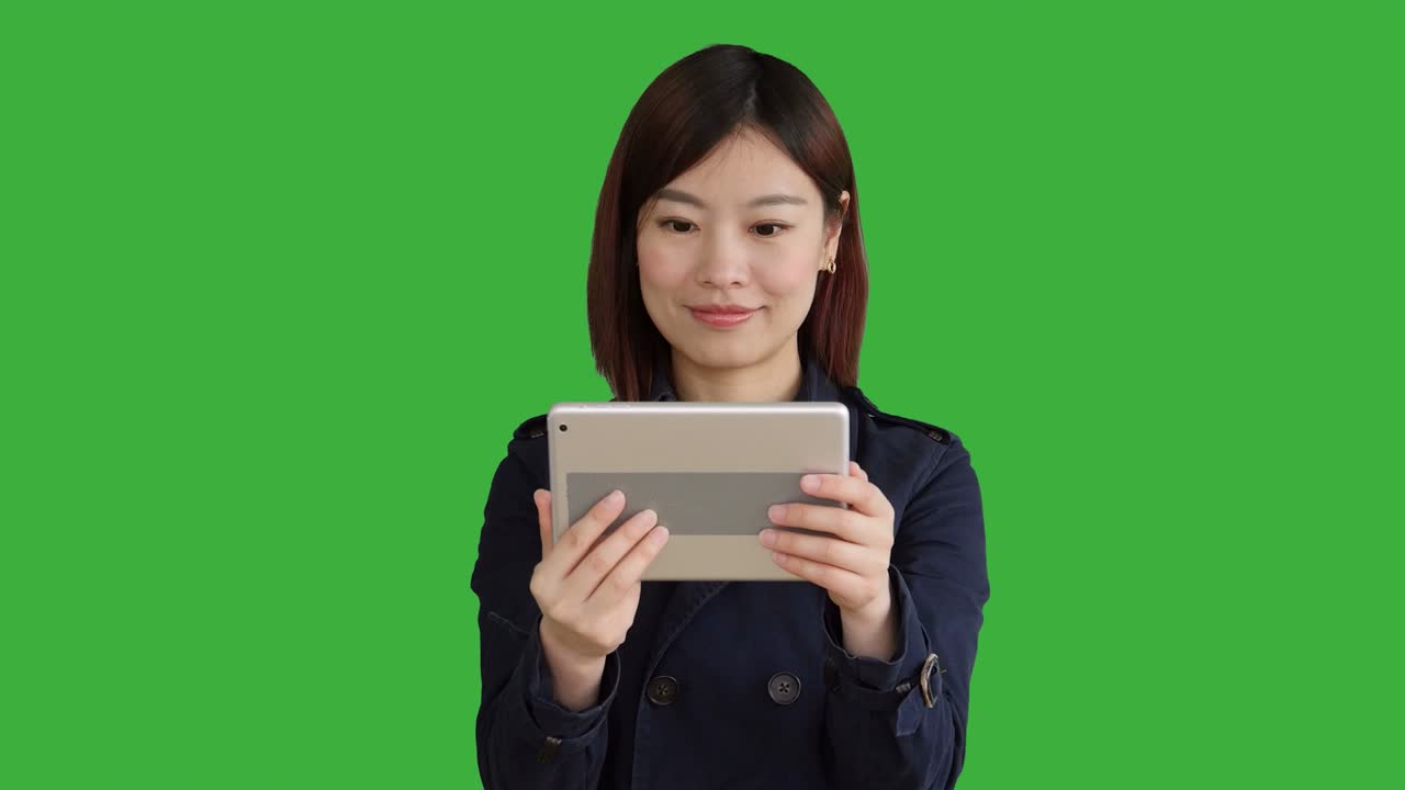 푸른 코트를 입은 젊은 아시아 여성, 스마트폰, 디지털 태블릿을 사용하고 카메라를 보고, 크로마 키를 위해 녹색 화면 앞에 서있는 여성.