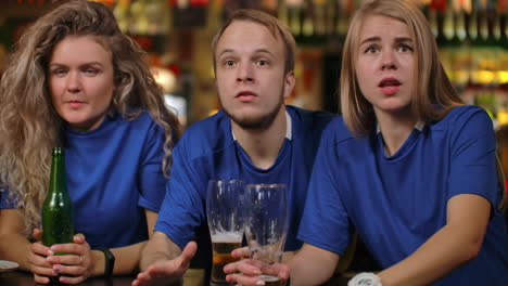 close-up group of fans emotionally watching a sports broadcast in a bar on tv with beer men and women in blue t-shirts of different races are upset after losing and losing their team.