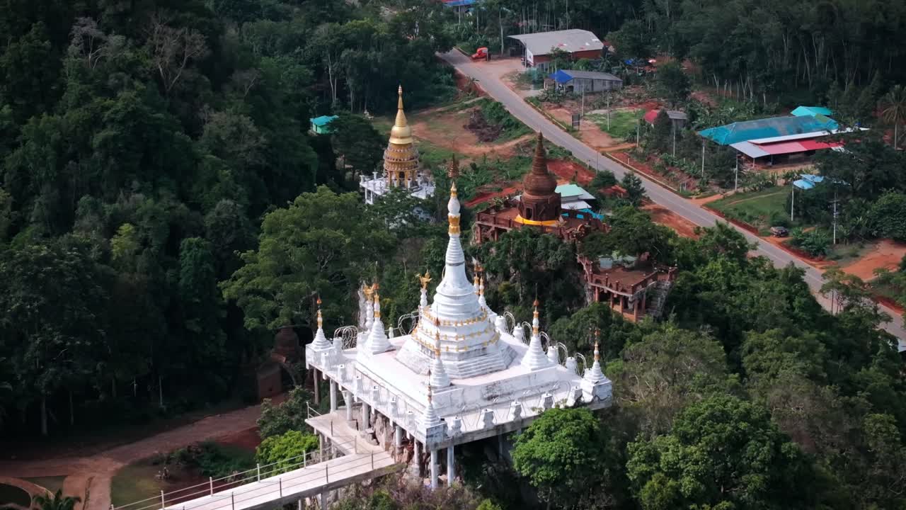 Drone footage of ornate hilltop Buddhist temples surrounded by tropical forest and a rural road in Southern Thailand. Aerial fly-away shows peaceful scenery