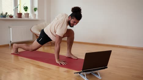 Active bearded man doing yoga following instruction video online trainer on laptop at home