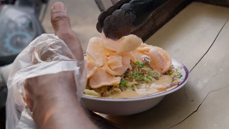 indonesian street food vendor adding shrimp crisps to savory porridge, close-up