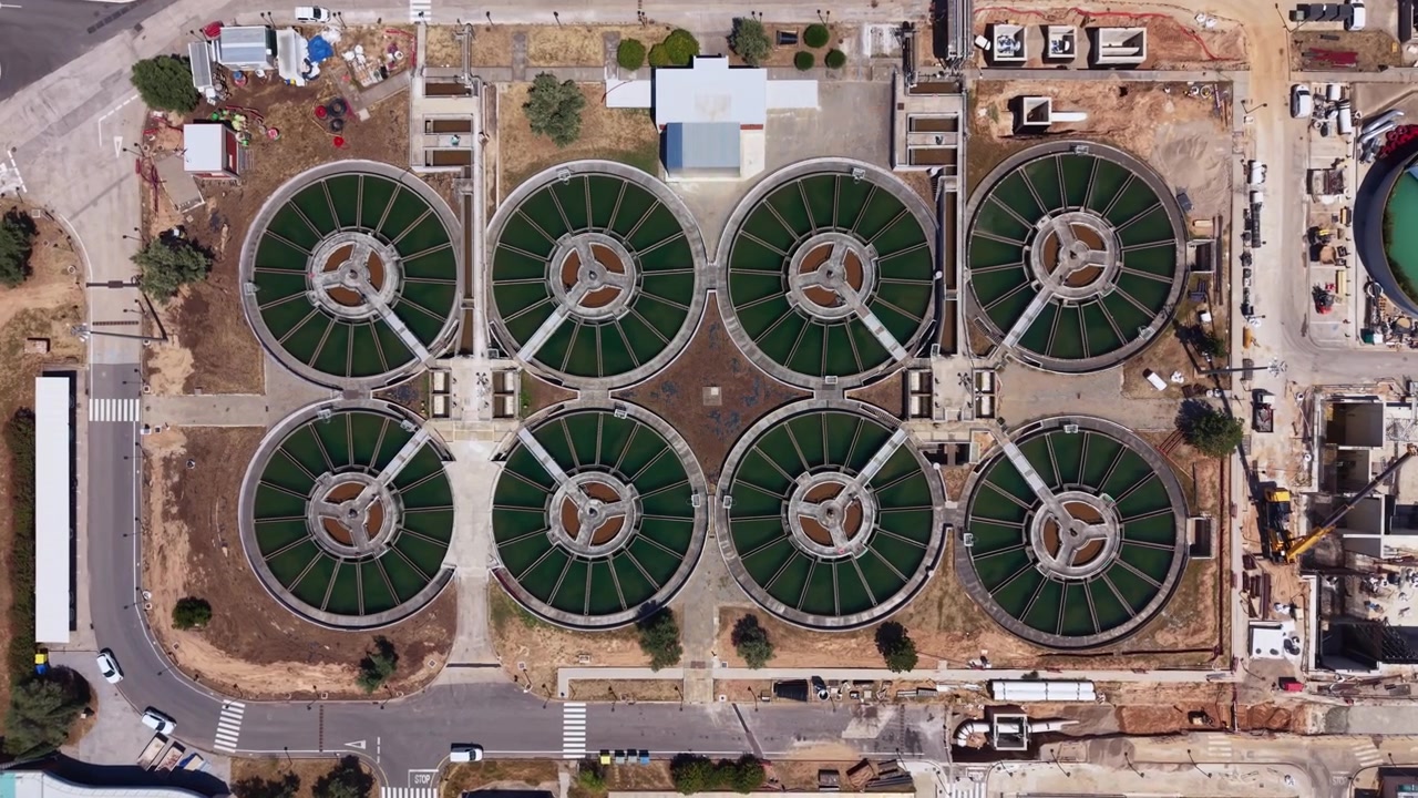 Top-down view of a water treatment plant featuring eight large circular clarifier tanks arranged symmetrically, surrounded by construction activity and urban infrastructure