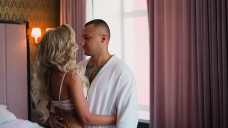 man hugs blonde lady in lacy bra standing in hotel room