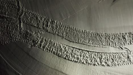 drone shot descends onto a snow groomer climbing a slope at night, revealing fresh tracks in snowy andorran mountains.