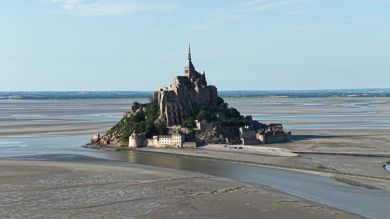 Filmisk droneudsigt fra luften af Mont Saint-Michel, der rejser sig over de vidtstrakte vadehavsområder ved lavvande. Det ikoniske middelalderkloster, den befæstede ø og snoede tidevandskanaler.