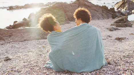 rear view of african american couple wrapped in blanket sitting on the rocks near the sea