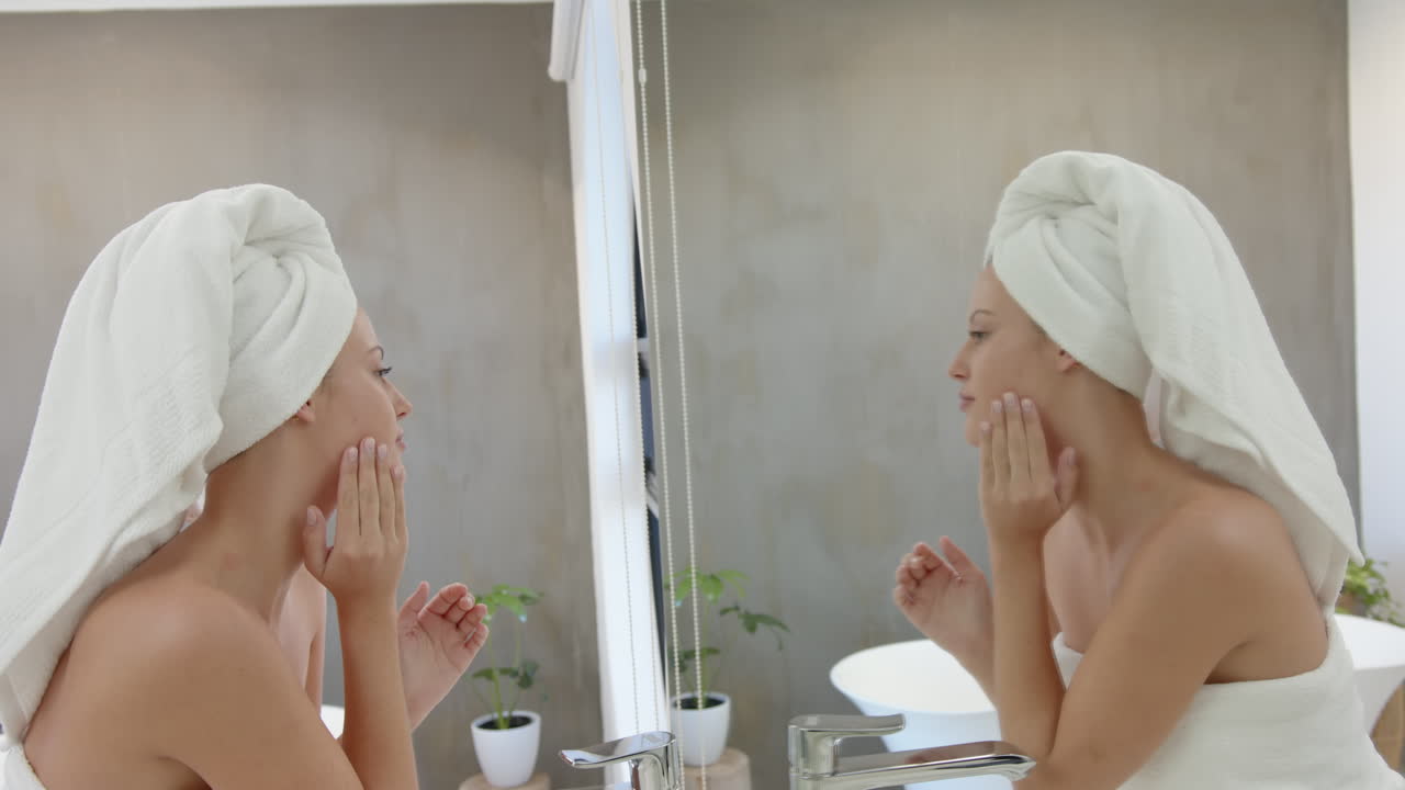 Looking in mirror, woman with towel on head touching her face in bathroom
