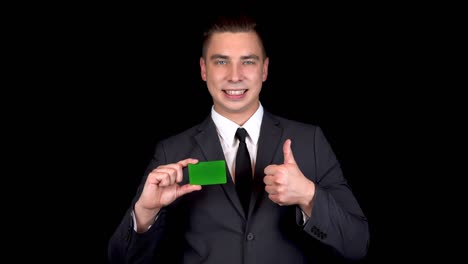 joven empresario presenta una tarjeta verde del banco y muestra como con su mano. tarjeta verde chromakey. hombre en un traje negro sobre un fondo negro