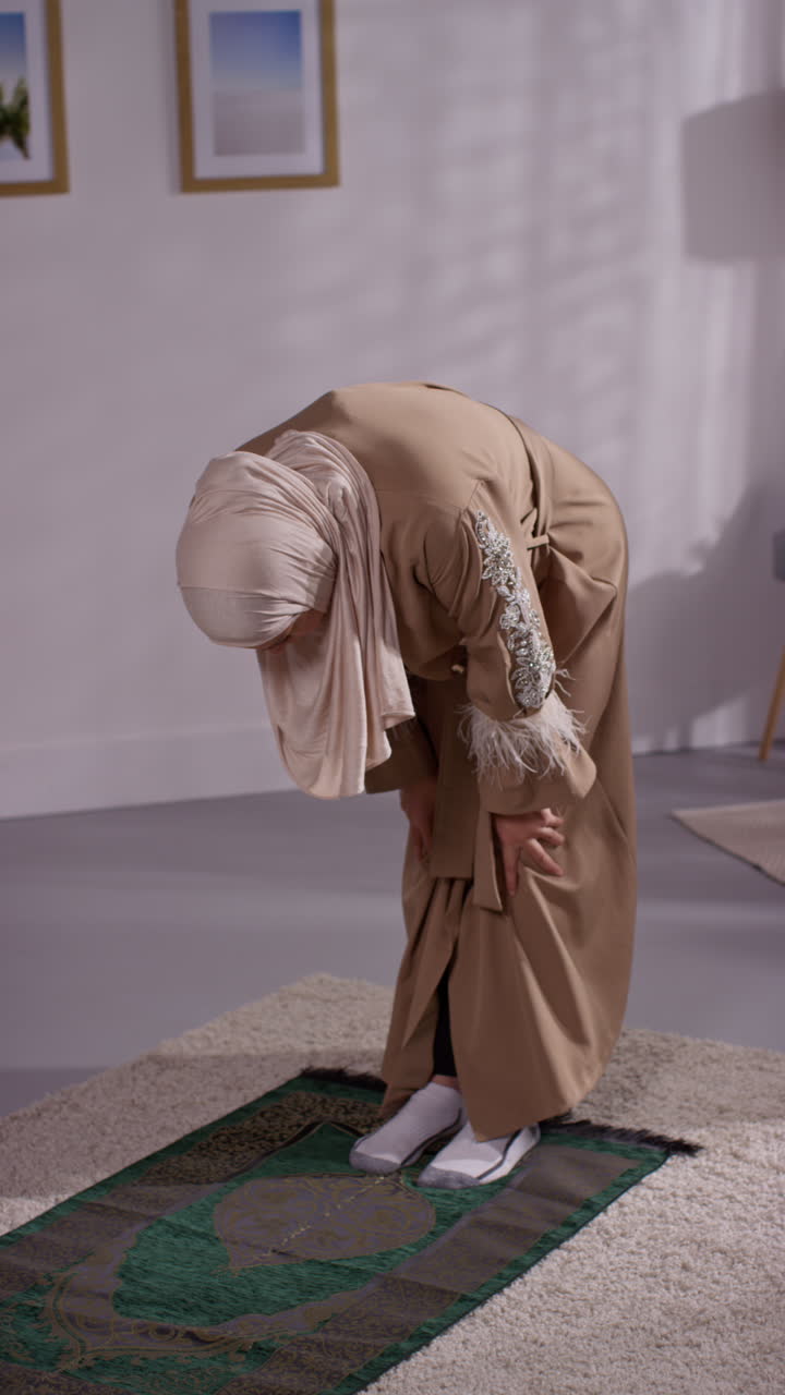 Vertikal video av muslimsk kvinne med hijab som ber knelende på bønneteppe hjemme 3