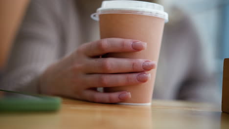 close-up hand view of lady tapping coffee cup on wooden table with partial view of phone in background, creating a cozy atmosphere with a soft blur