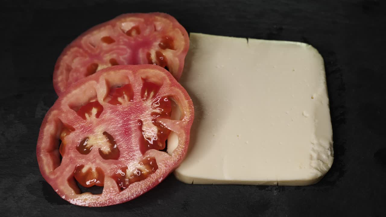 A close-up overhead shot of two slices of tomato and a block of fresh cheese placed on a dark surface, while extra virgin olive oil is slowly poured over them