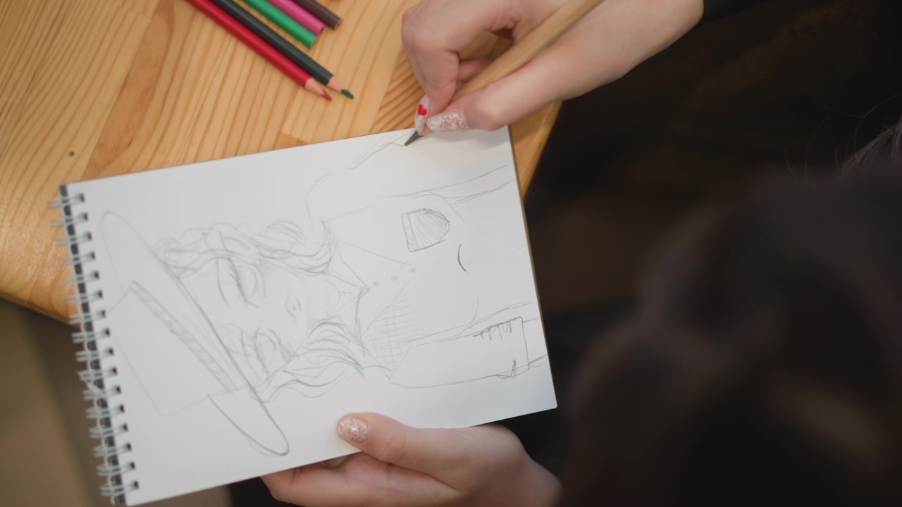 Top down view of person sketching in spiral bound notebook, right hand guiding pencil across page while left hand steadies paper on wooden table in bright indoor workspace, with drawing, sketchbook