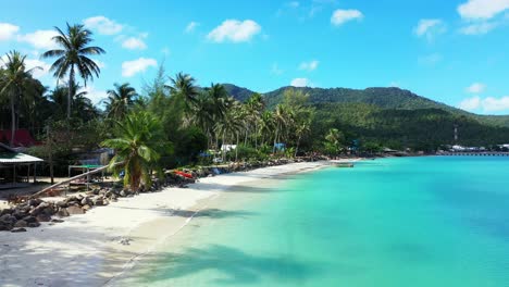 Paradise-beach-with-palm-trees-bent-over-white-sand-washed-by-calm-clear-water-of-turquoise-lagoon-on-bright-blue-sky-with-clouds-background-in-Thailand