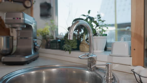 close up of shiny chrome kitchen faucet near stainless sink with electric blender machine in background, potted plants on windowsill, and eggs arranged neatly in white egg rack beside soap dispenser
