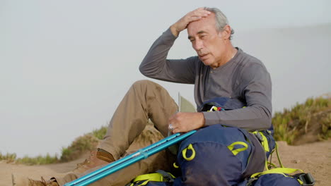 medium shot of a tired hard-breathing hiker sitting on ground, taking off cap and wiping sweat from forehead