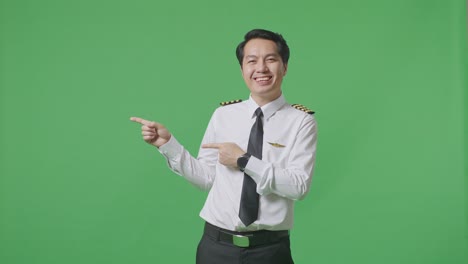 아시아 남자 조종사가 미소 짓고 녹색 스크린 배경 스튜디오에 서서 을 가리키고 있습니다.