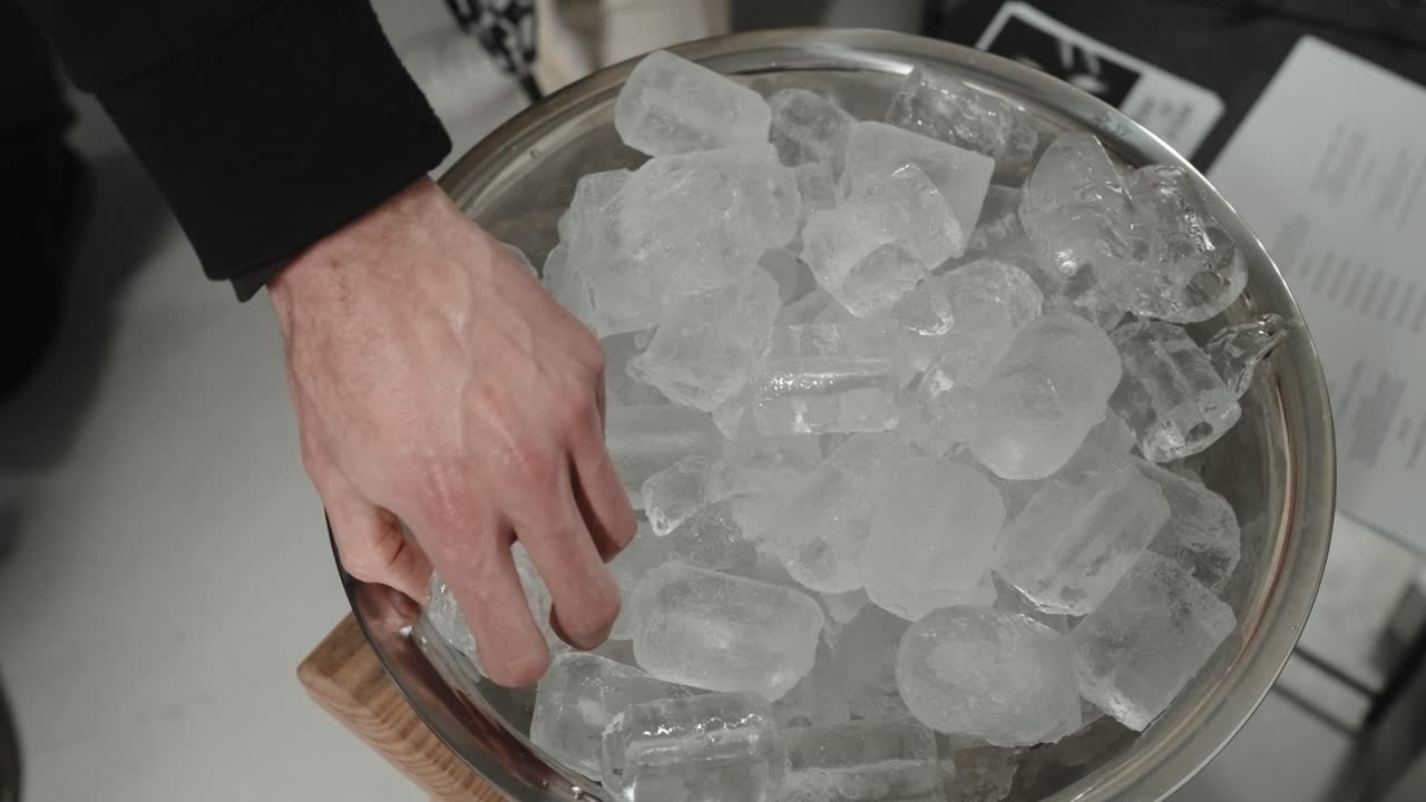 Primer plano de una mano seleccionando cubitos de hielo de un recipiente metálico sobre una mesa
