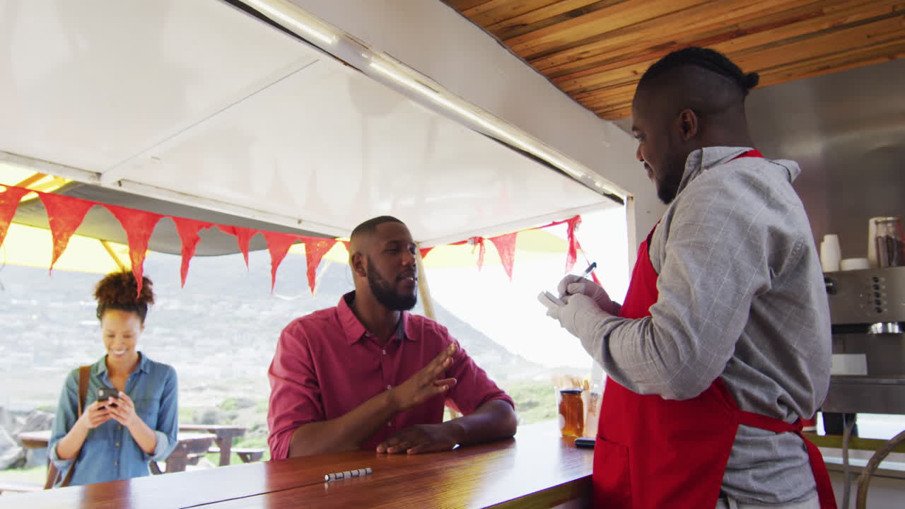 미소 짓는 아프리카계 미국인 남성 음식 트럭 소유자가 남자로부터 주문을 받고, 여성이 전화를 사용하여 기다리고 있습니다.