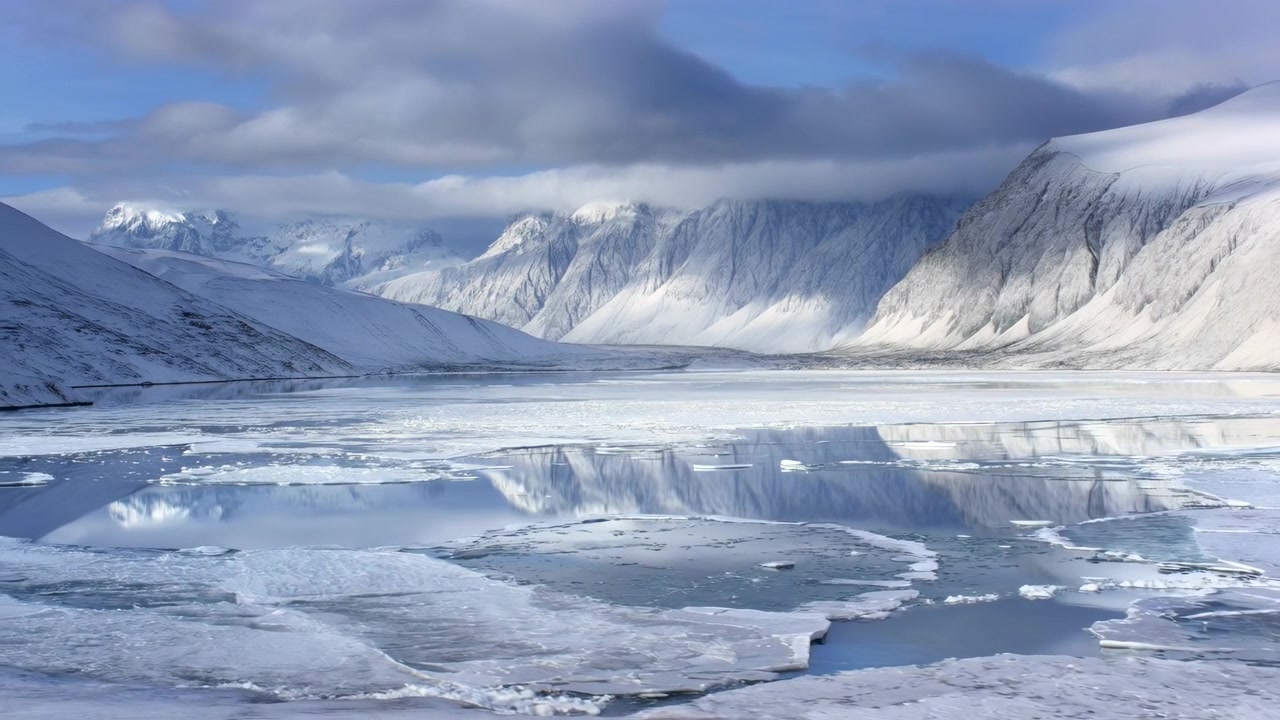 Brudte isflager driver hen over en frossen arktisk fjord, mens åbne render udvides og afslører reflekterende mørkt vand ved siden af snedækkede bjerge under et lavt skydække og roligt vinterlys