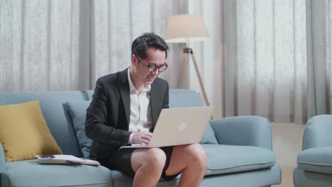 소파에 앉아서 집에서 노트북으로 타이핑하는 재과 쇼트를 입은 미소 짓는 아시아 사업가