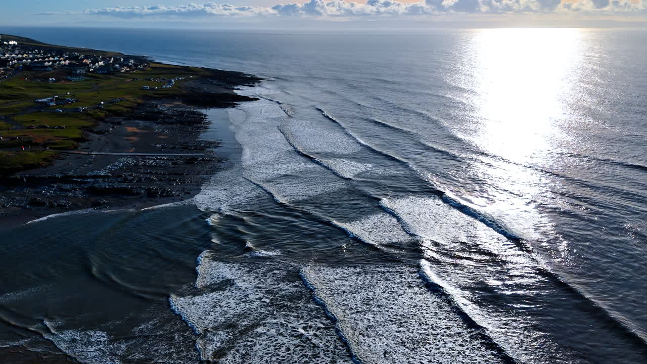 Høyt vinkelbilde av havbølger som bryter på stranden. Sollys reflekteres på sjøvannet med bølger som ruller inn mot en mørk steinete og sandete kystlinje på dagtid