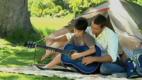 padre enseñando guitarra a su hijo frente a una tienda de campaña