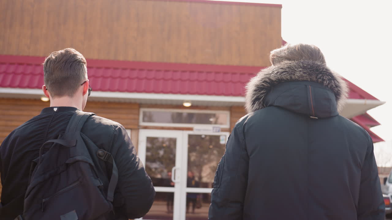 Two young men in winter jackets stand facing building entrance with red roof, one carrying black backpack and other with fur hood, urban outdoor scene with bright daylight atmosphere and wooden facade background