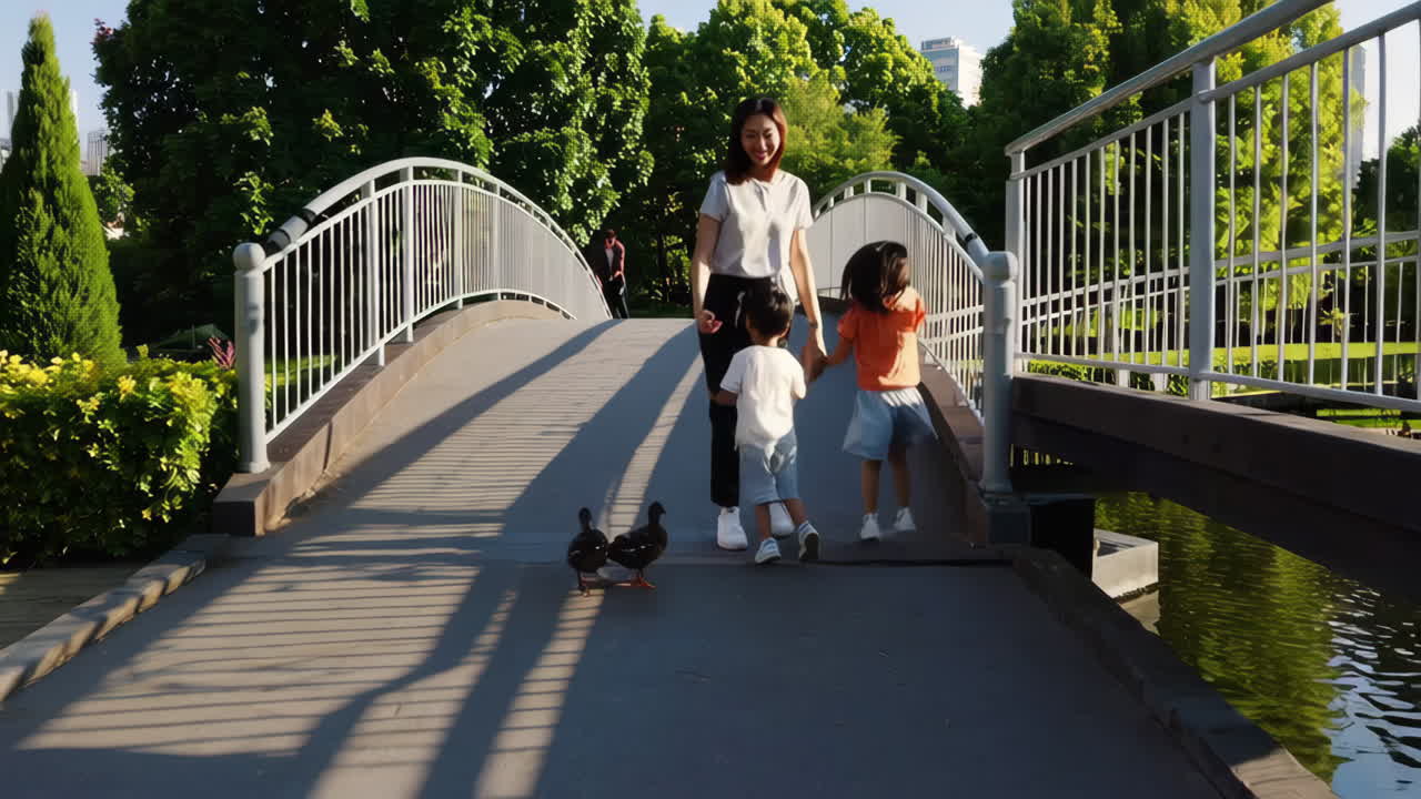 A family, consisting of a mother and her two children, walks hand-in-hand across a bridge in a sunny park, encountering a duck on their path.