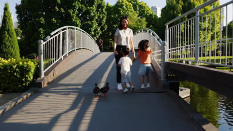 a family, consisting of a mother and her two children, walks hand-in-hand across a bridge in a sunny park, encountering a duck on their path.