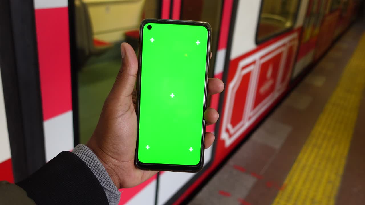 Person holding a smartphone with a green screen at a train station
