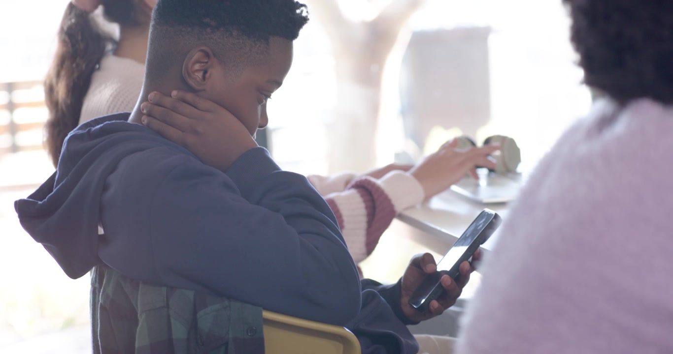 African American teens sitting at window counter boy tapping smartphone while girl moving small cup