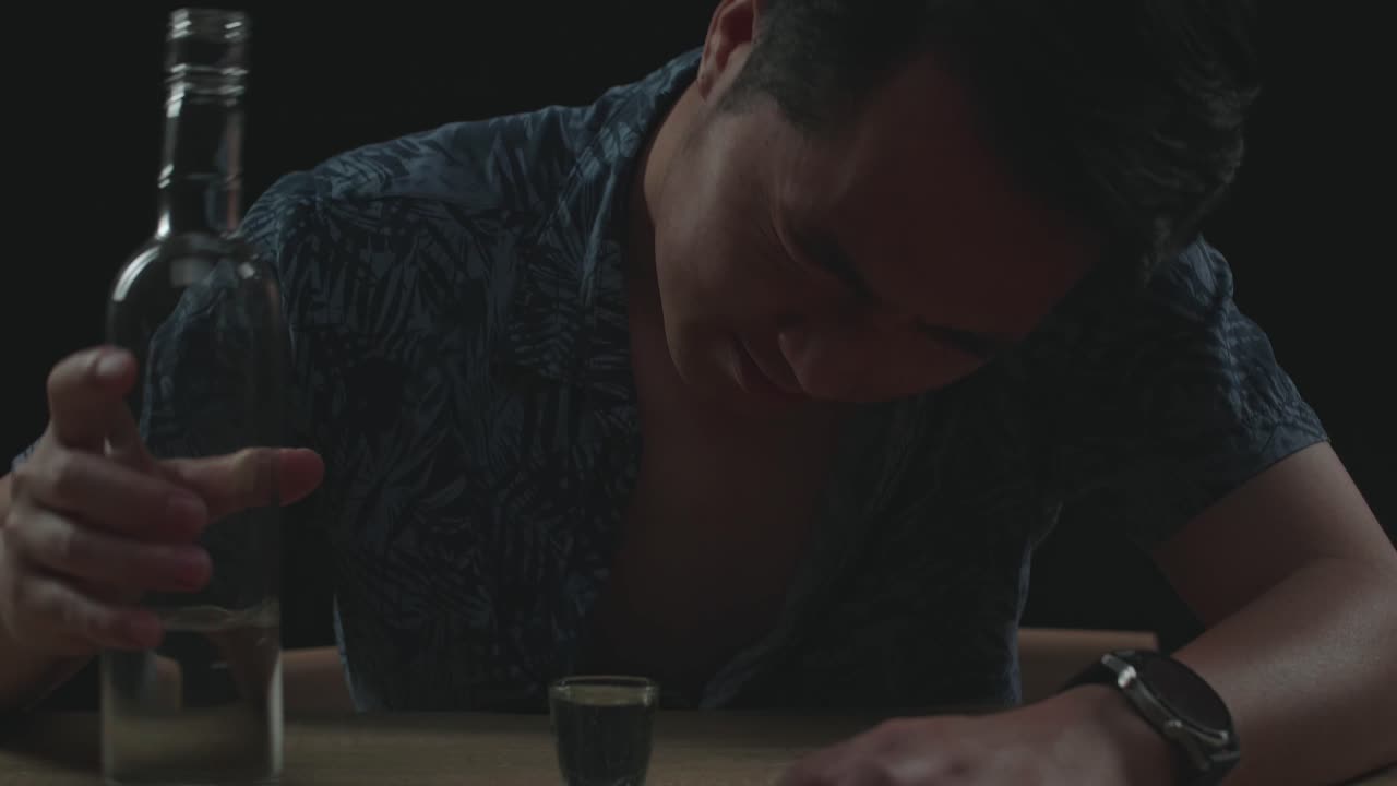 술에 취한 아시아인 남자가 검은색 배경에서 음료를 마시기 전에 카메라에 보드카 컵을 환호합니다.