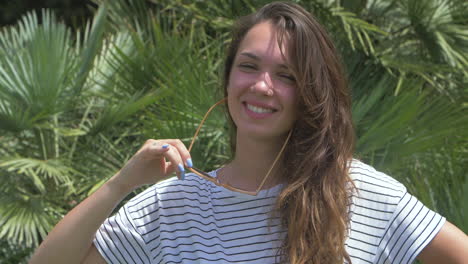 una mujer joven sonríe a la luz del sol, con gafas de sol y una camisa a rayas. el fondo es un exuberante paisaje tropical.