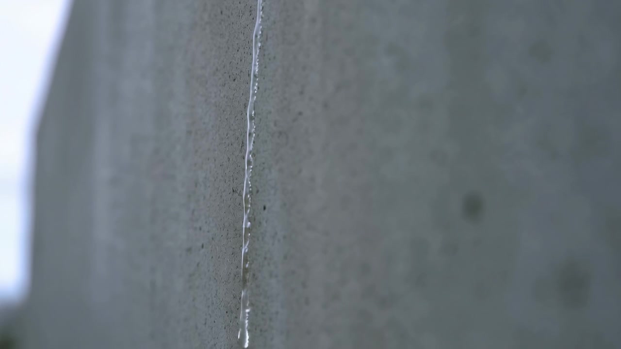 Movimento ravvicinato di una stretta scia d'acqua traslucida che aderisce all'esterno di cemento ruvido, concentrandosi in goccioline e una goccia pendente, mentre una striscia bagnata si allarga lentamente su una superficie testurizzata in una luce soffusa.