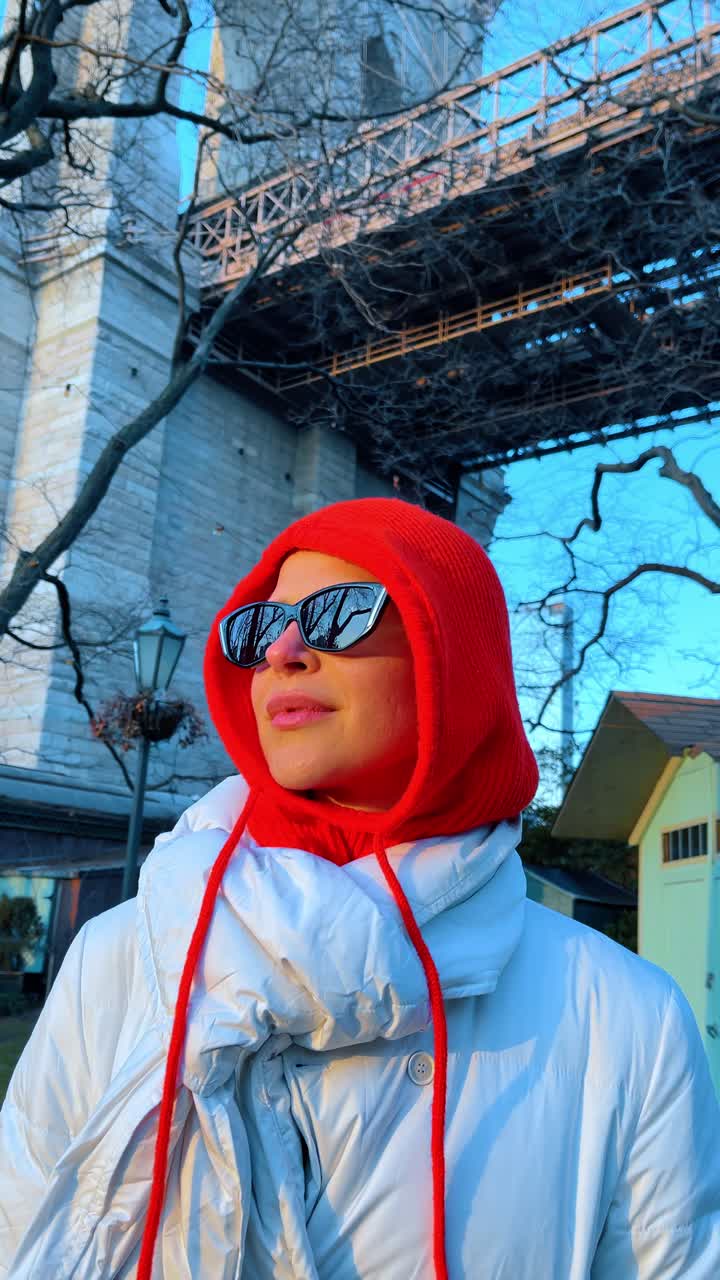Donna in piedi sotto la struttura del Ponte di Brooklyn. Donna con cappotto bianco e cappuccio rosso in piedi sotto il Ponte di Brooklyn con alberi invernali e torre in pietra.