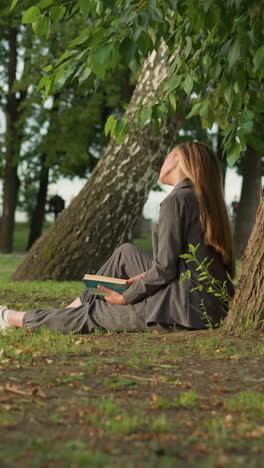 dama de ropa gris se sienta al aire libre bajo un árbol, leyendo un libro con la mano en la página, inclinando la cabeza y la pierna extendida, el fondo presenta vegetación y árboles